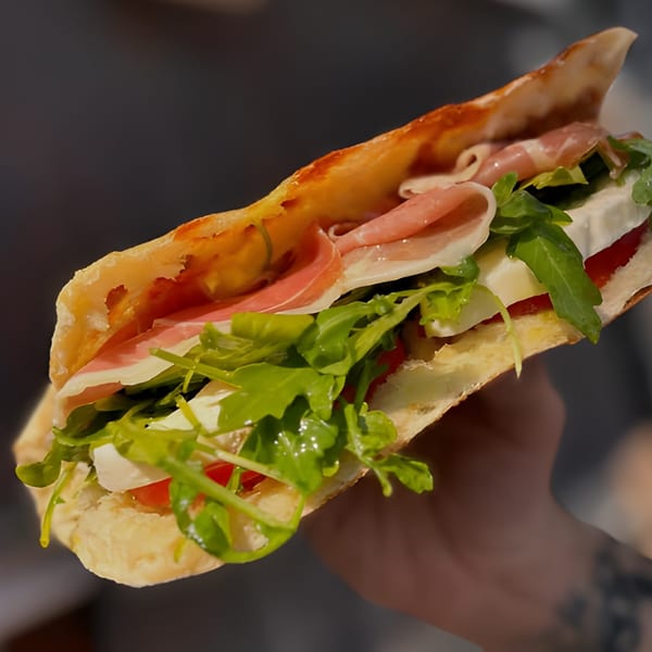Hand holding a Pan and Peel Pizza sandwich filled with prosciutto, mozzarella, arugula, and tomatoes.