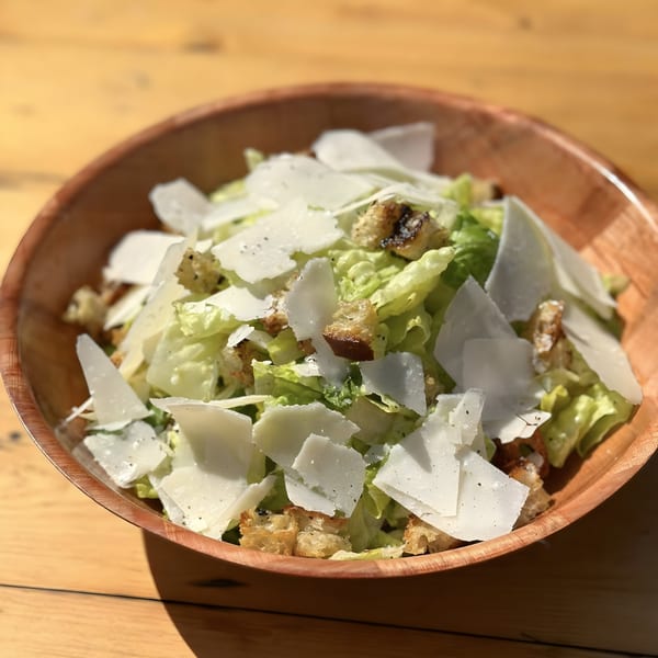 A wooden bowl filled with fresh Caesar salad, topped with large parmesan shavings and croutons.